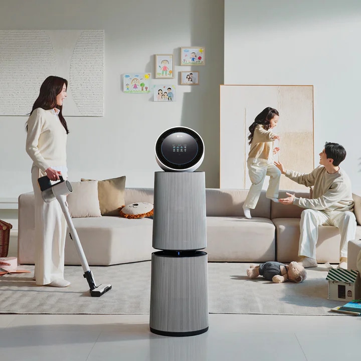 거실 공간에 LG 퓨리케어 AI 오브제컬렉션 360˚ 공기청정기가 있습니다. 소파에 앉아 있는 남성과 뛰어노는 아이, 그리고 청소기를 들고 있는 여성이 보입니다. 벽에는 그림이 걸려 있고, 바닥에는 화분과 장난감 등이 배치되어 있습니다.